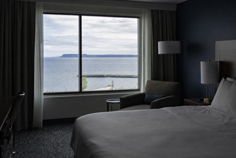 A hotel room with a made bed and an armchair by a large window over a wide lake and a low peninsula