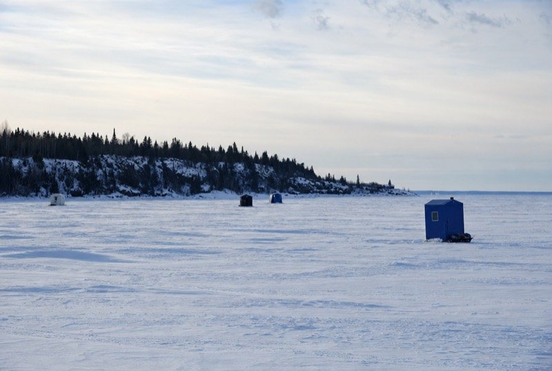Small ice-fishing shelters spread across a snow-covered frozen bay with dark spruce along the shore